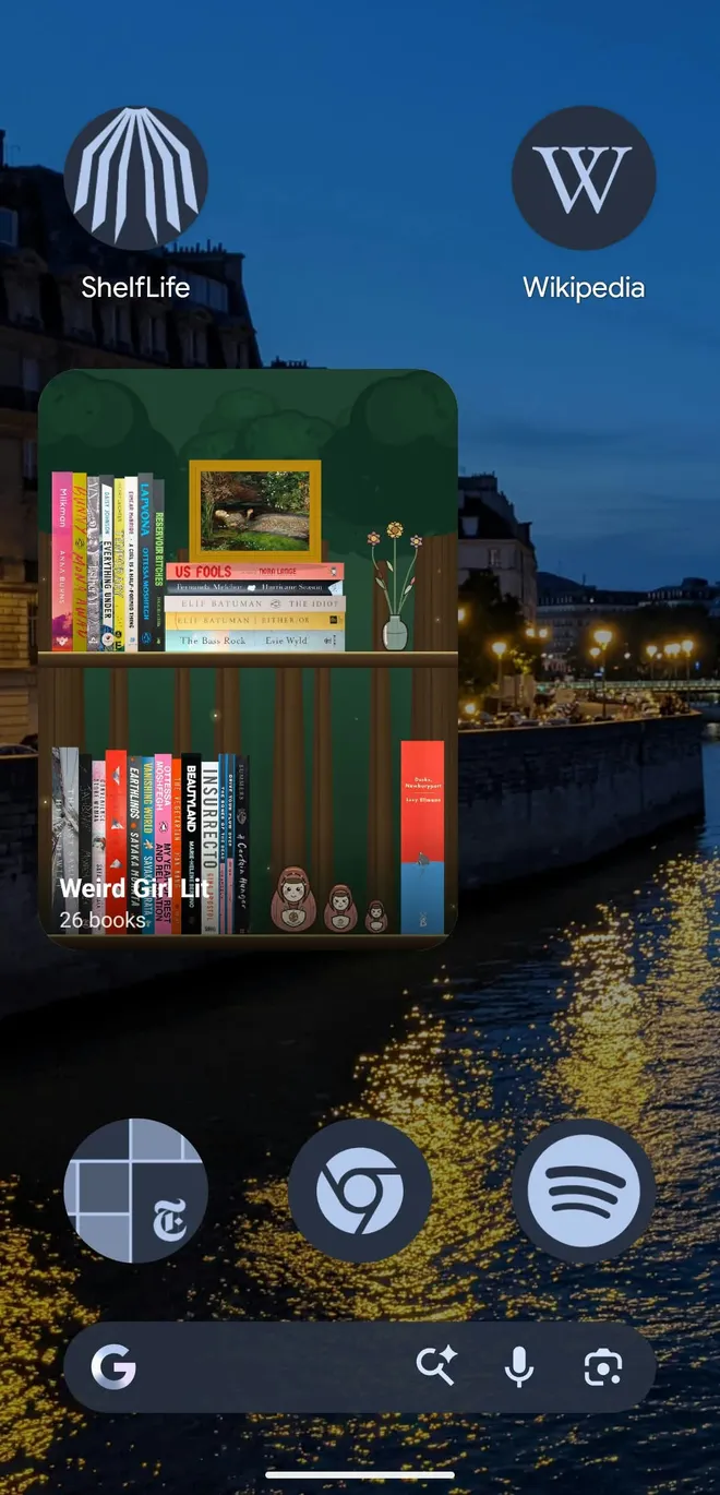Android home screen with a tall ShelfLife widget showing a shelf named Weird Girl Lit, 26 books, on two rows against a forest backdrop with a framed painting, a flower vase, and nesting dolls